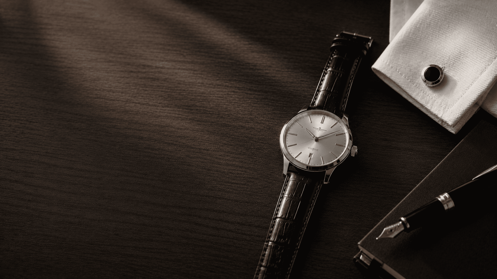 Slim dress watch on a dark wood desk with a folded shirt cuff